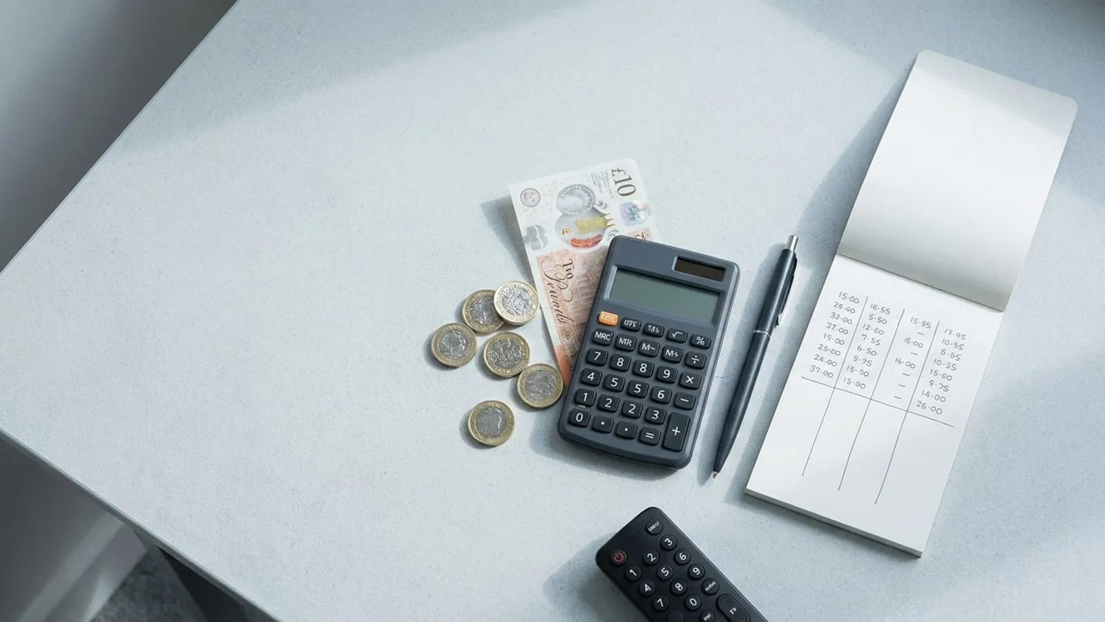Pound coins, a calculator and a notepad of figures laid out on a desk beside a TV remote