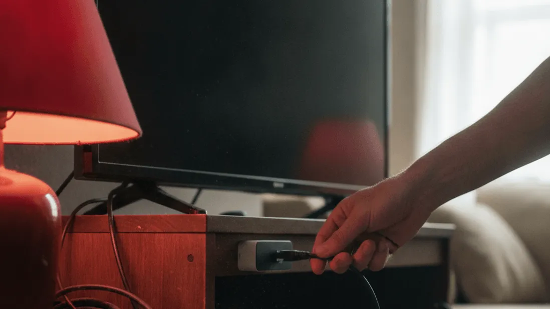A hand plugs an ethernet cable into a small box behind a television stand, with cables and warm lamplight.