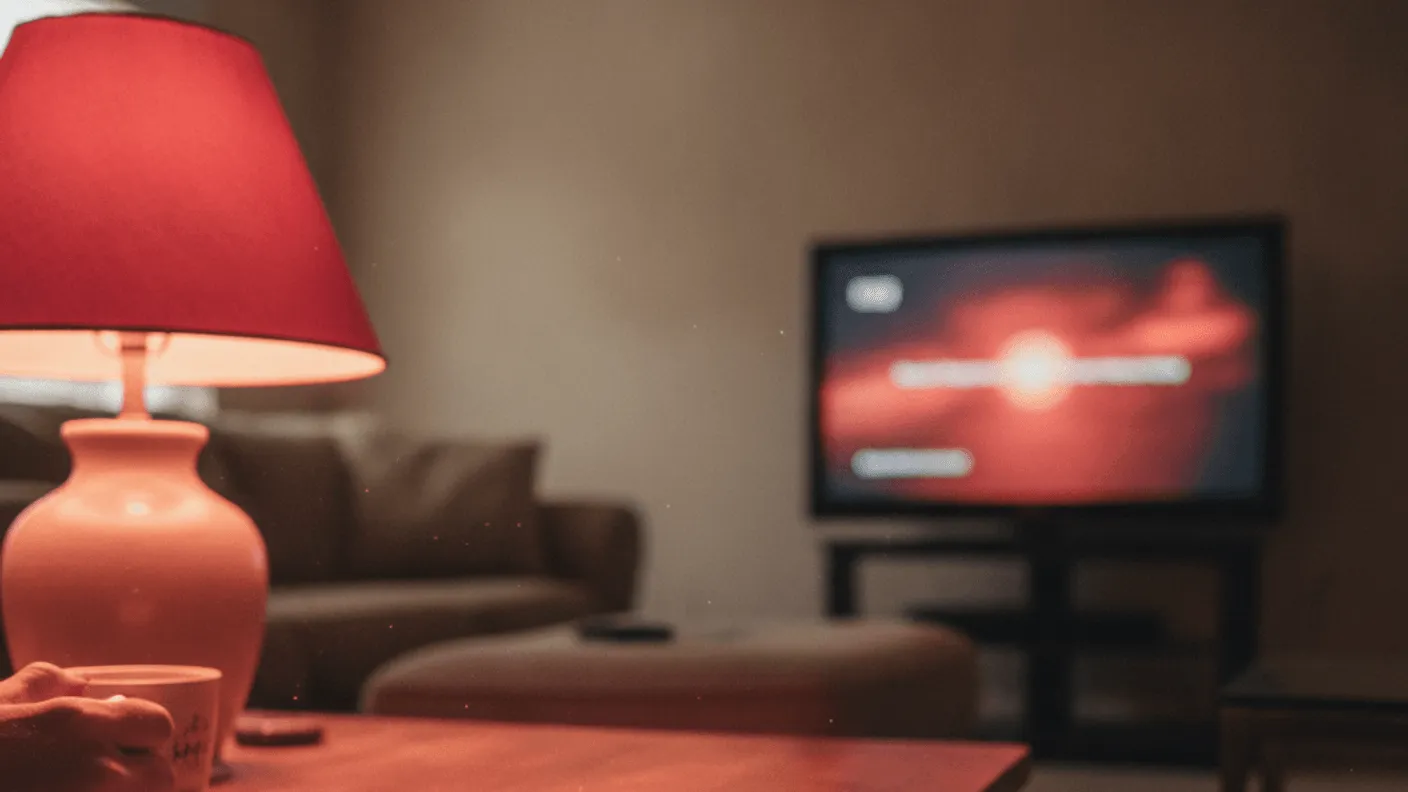 A remote and a folded paper bill on a coffee table in evening light, with a television in the background.