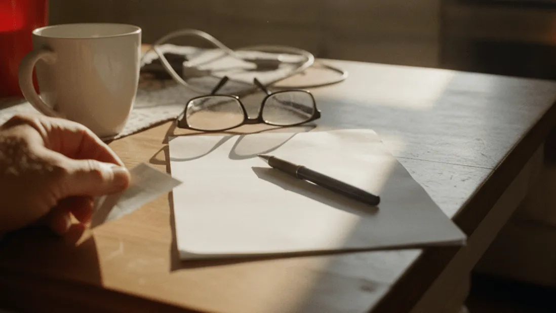 A hand reaches for a blank bill on a kitchen table, next to reading glasses, a pen, and a coffee cup.