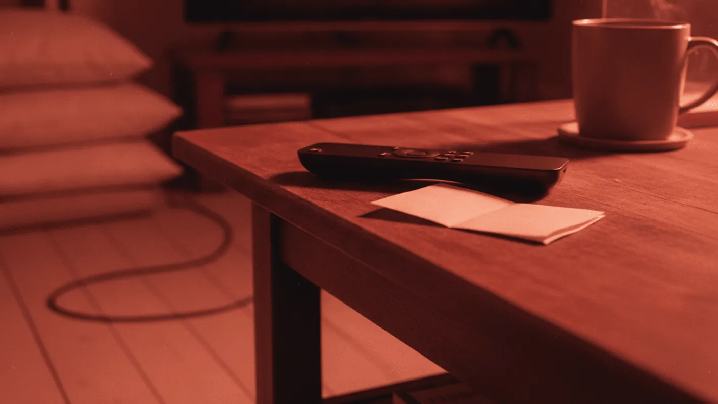 A remote control and folded paper rest on a wooden coffee table, with a dark television screen in the background.