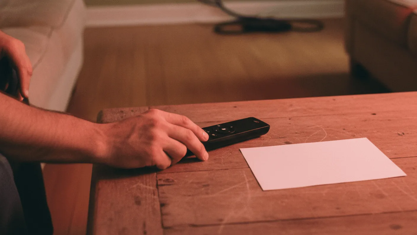 A hand reaches for a remote control on a coffee table beside a paper bill, with a television glowing in the background.