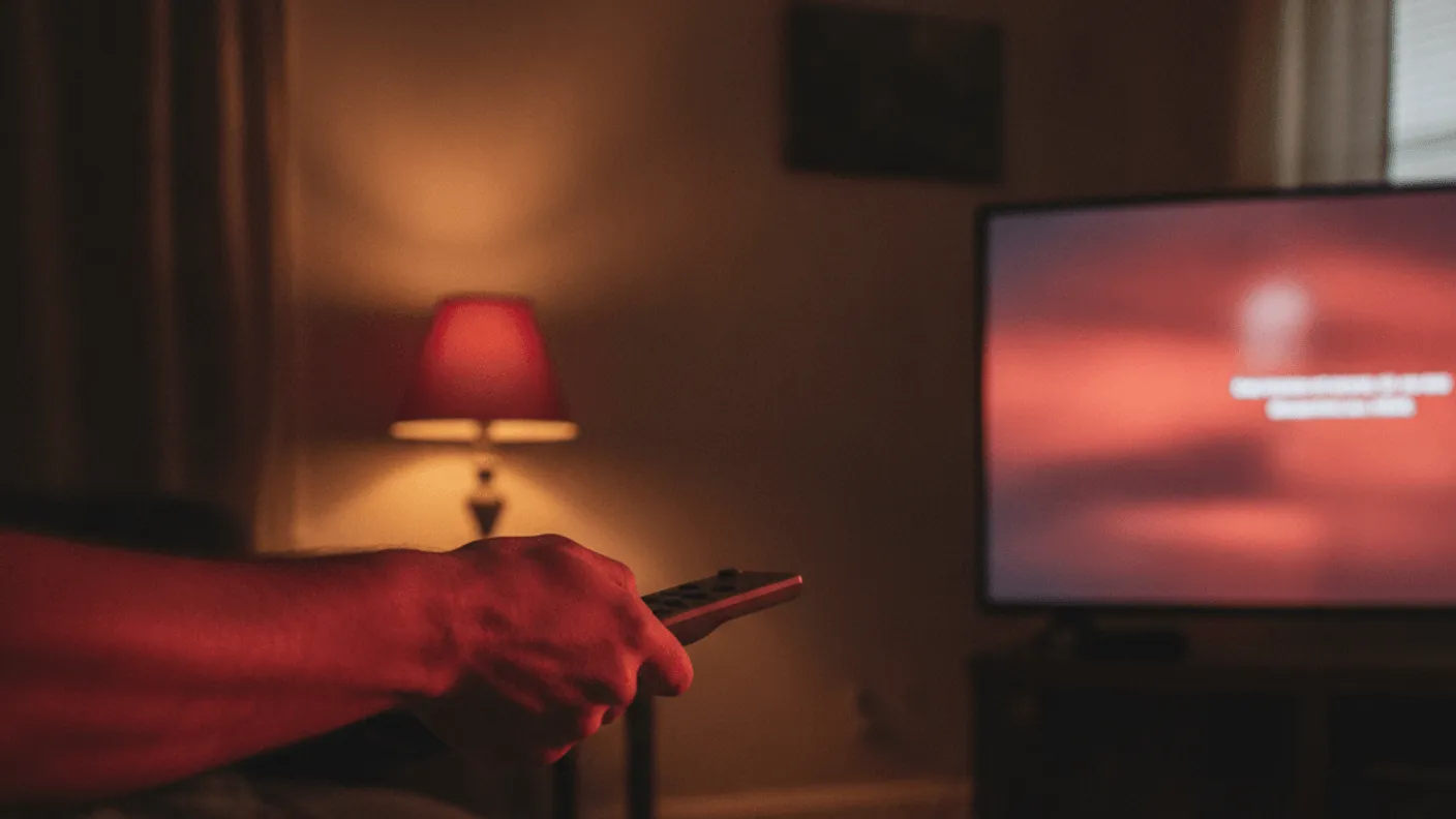A hand holds a remote, pointing it toward a television screen glowing red in a dark living room.