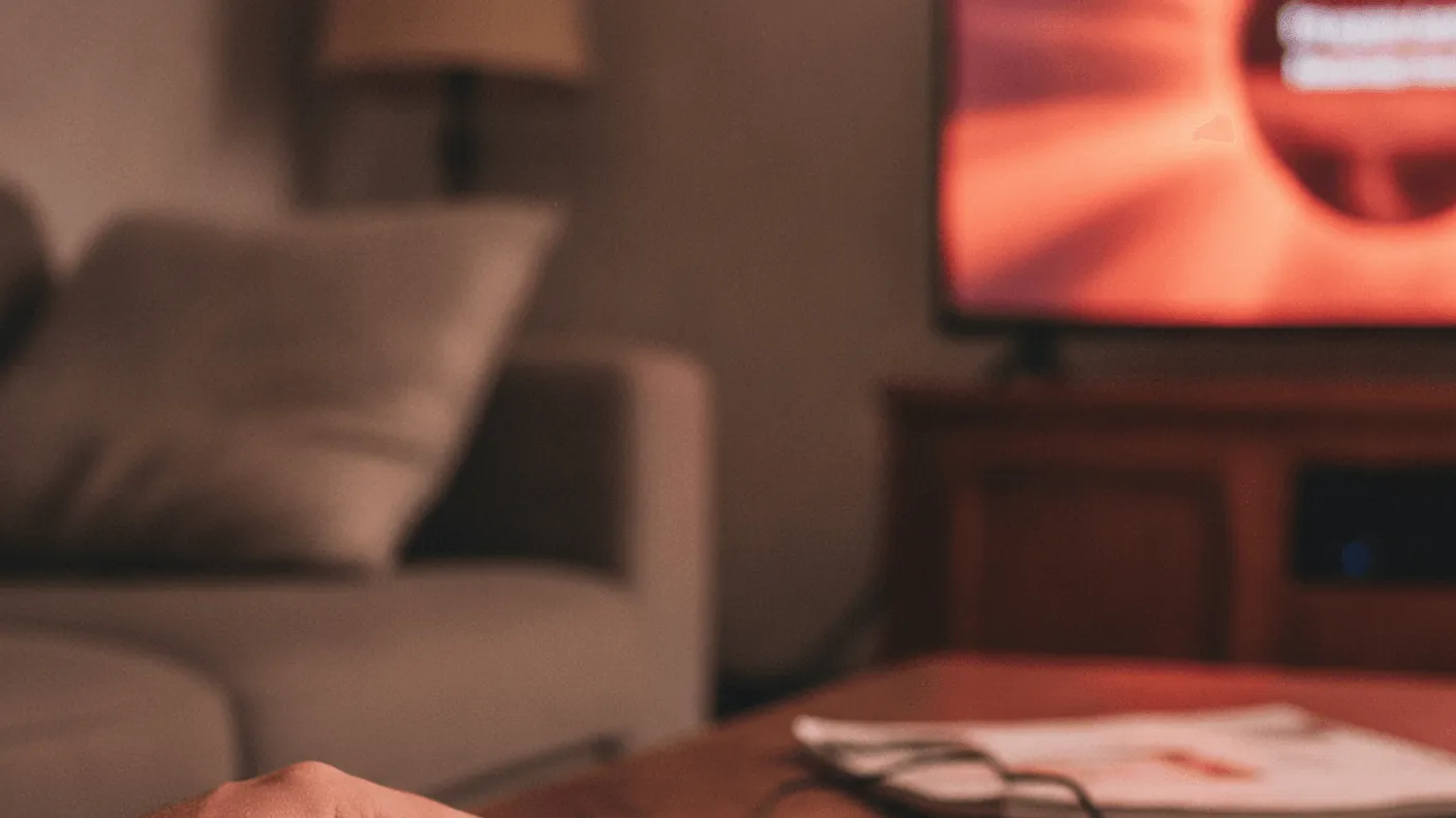 A hand reaching for a remote control and a small streaming device on a wooden coffee table in a dim room.